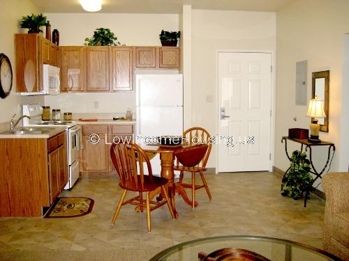 Interior of a kitchen equipped with a gas range, a twin sink, a refrigerator/freezer and several storage cabinets a small table and two chairs.