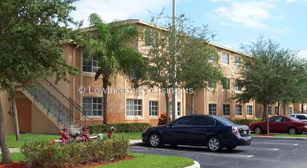 Large residential apartment block with red brick construction.  Exterior stairway to second floor. Large windows facing on in street parking. 