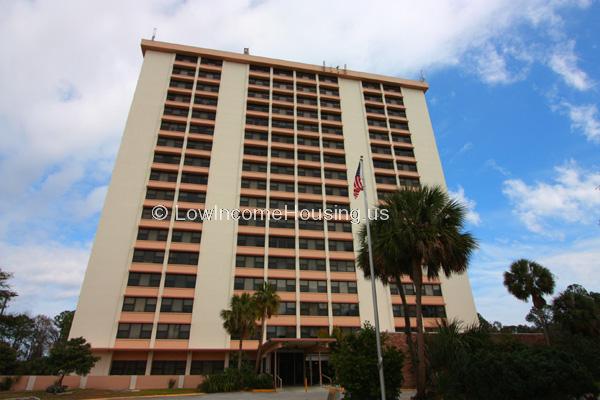 This is a photograph of a large apartment building (17 story with 8 dormer units.  Building is located in area with ample foliage.  