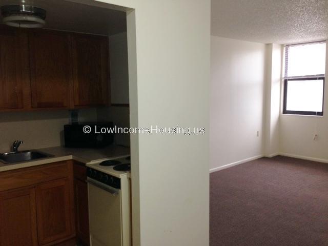 Interior photograph of apartment with partial view of kitchen and view of bedroom with window to exterior. 