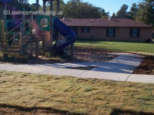 Large red brick building located behind a playground equipped with a slide and climbing equipment.   