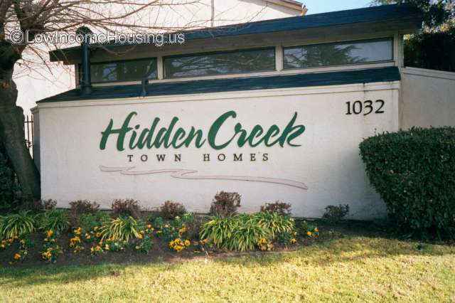 Picture of an outdoor wall with the name Hidden Creek Town Homes and the address number 1032. Three windows, some grass, bushes, and flowers.