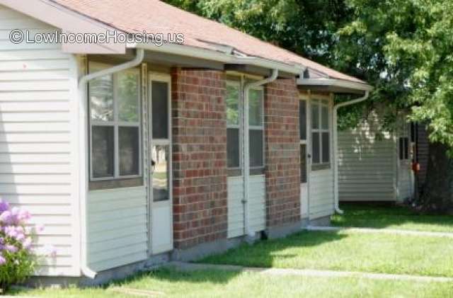 This is a photograph of a small wooden framed house with two entrance doors and 4 windows.  The roofing is shingles and there is a metal gutter attached to the roof. 