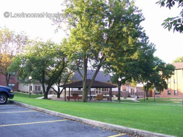 Photograph of several (5) mature trees that frame  a photograph of a covered area that appear to be chairs and tables.