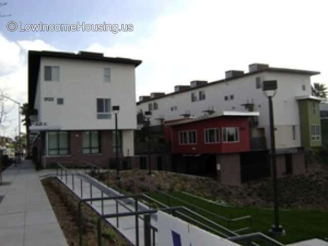 Handicap ramp with two  white three story buildings in the back with red and green accent walls. Sidewalk on the left, green area to the right and lamp posts.