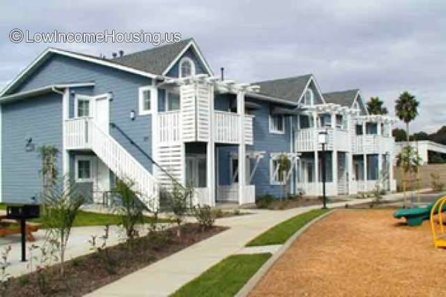 Three two story buildings w/ terraces and exterior staircases facing playground and sidewalk with well kept landscaping.