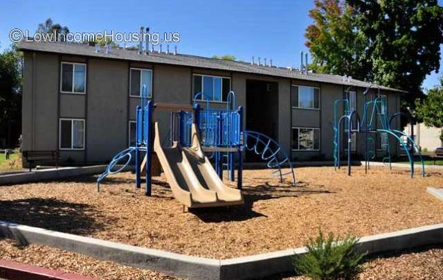 Two story Red Brick town houses with playground equipment surrounded by wooden barrier for children.  Towne houses are equipped with large picture windows facing playground.  