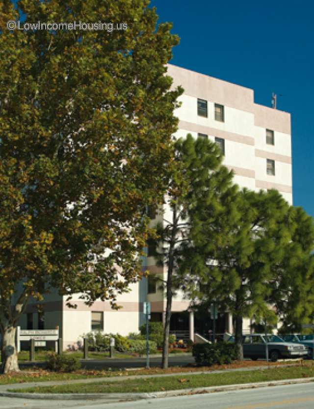 Concrete and brick construction of a housing unit with 8 window units, full access entrance facing street on ground floor.  Ample foliage provides shade.