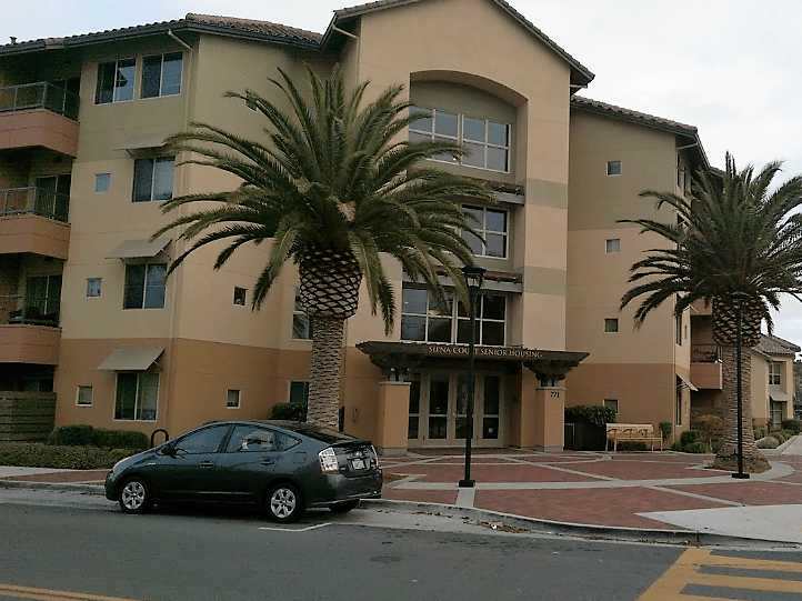 Corner streetview of four story building. Two large palm trees and light posts. a Toyota Prius is parked on the street. A bench is to the right of the entrance.