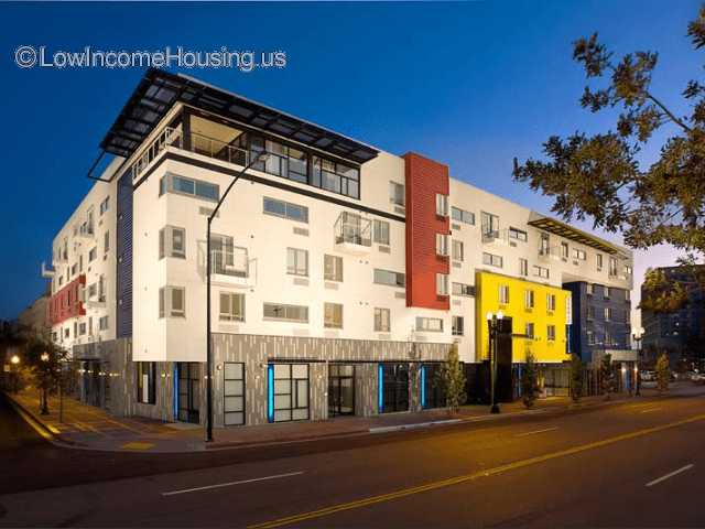 Corner shot of modern five-story residential building. Building has a balcony on the top corner red, yellow, and blue accent walls over white. Picture taken in the early evening.